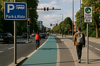 A modern urban road features a dedicated turquoise cycle lane with a cyclist in a red jacket riding away from the camera. To the right, a pedestrian with a grey backpack walks beside the lane on a clean pavement. Clear signage includes a “Park & Ride” sign and a “Clean Air Zone End” sign. The background shows light traffic, smart traffic lights, and tree-lined streets under a blue sky, representing eco-friendly city transport and smart urban planning.