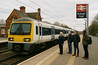 A high-resolution photo of a British railway station shows a modern white and blue train with a yellow front arriving at the platform. Five passengers of mixed age and background stand waiting beside a classic red-brick station building under overcast skies. A vintage-style British Rail sign is prominently displayed. The image represents rail renationalisation, public ownership, and investment in accessible, community-focused transport infrastructure.