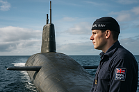 A Royal Navy sailor stands on the deck of a British Vanguard-class nuclear submarine in open waters, wearing a navy uniform with a visible Union Jack and “Royal Navy” patch. The image symbolizes the UK’s nuclear deterrent, maritime defence, and strategic military readiness at sea.