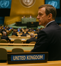 A British delegate stands inside the United Nations General Assembly hall, with the “UNITED KINGDOM” nameplate in focus. The man observes the session against a backdrop of golden walls and UN insignia, symbolizing the UK’s reassessment of global alliances and multilateral commitments.