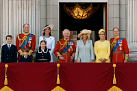 A photograph of the British Royal Family shows them gathered on the balcony of Buckingham Palace during a ceremonial event. Dressed in formal attire, all family members—including King Charles III, Queen Camilla, Prince William, Princess Catherine, their children, and other senior royals—face the camera with composed expressions. Ornate columns and velvet banners frame the scene, capturing a dignified and modern image of the monarchy.