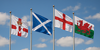 A high-resolution photograph taken around midday on a clear, partly cloudy day shows four national flags of the United Kingdom flying proudly on separate silver flagpoles. From left to right: the Ulster Banner of Northern Ireland, the Saltire of Scotland, the St. George’s Cross of England, and the Red Dragon of Wales. The flags wave vibrantly against a bright blue sky, representing unity and equal national identity across the UK.