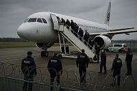 Photograph of a deportation operation at a UK airport, showing immigration enforcement officers escorting individuals up a mobile stairway into a white aircraft. The area is surrounded by secure fencing with barbed wire, and a marked UK Immigration Enforcement van is visible nearby. The sky is overcast, reinforcing the serious and controlled tone of the operation.